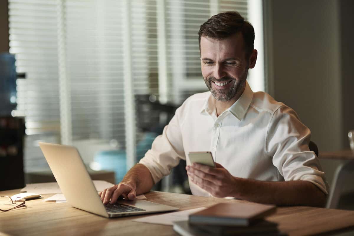 Corretor de seguros sorrindo enquanto usa o celular e o notebook para gerenciar o Instagram da corretora