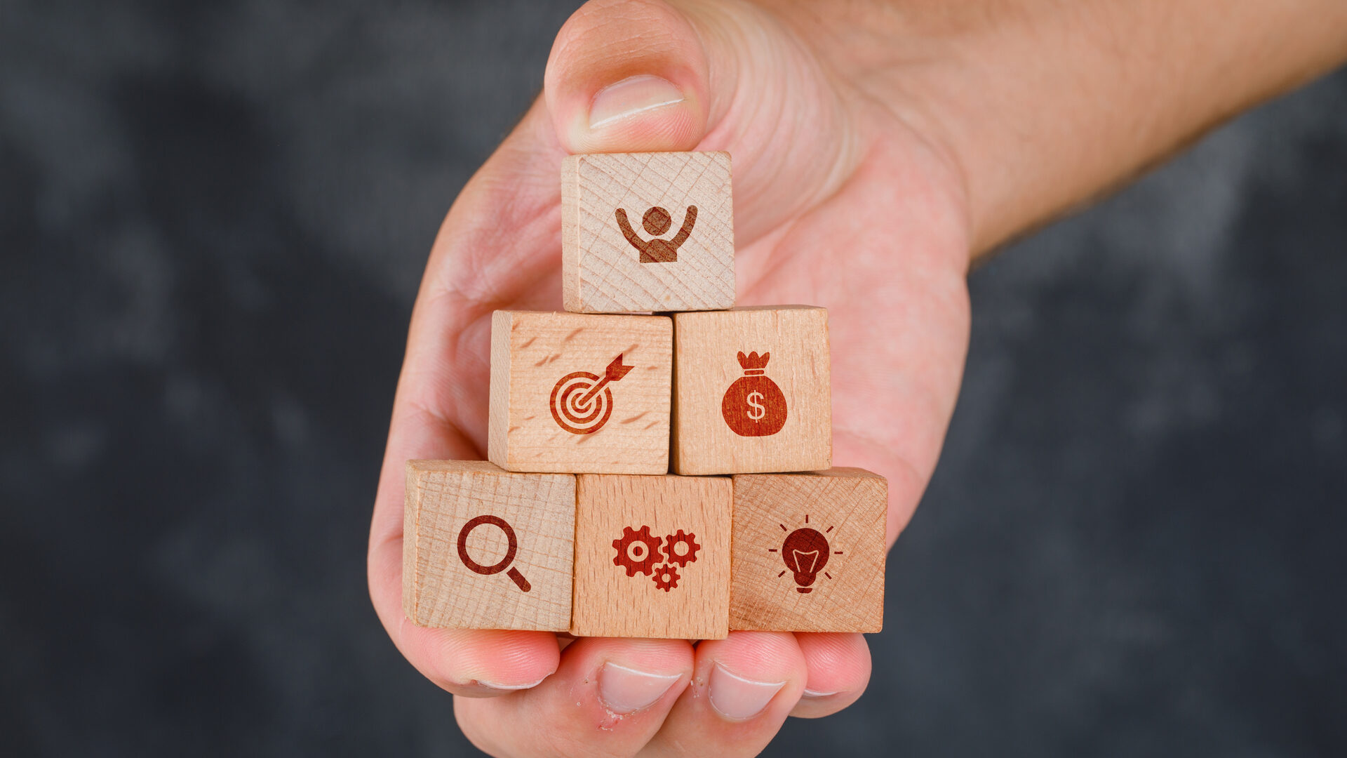 Mão segurando blocos de madeira com ícones de metas, dinheiro e ideias — soft skills corretor de seguros